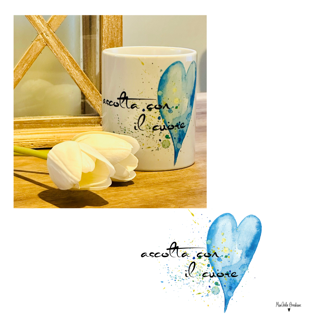 Tazza in ceramica bianca con grafica colorata con un cuore blu nei toni pastello e frase motivazionale "Ascolta con il cuorei", appoggiata su tavolo di legno con fiore decorativo e cornice sullo sfondo. Design originale Monjulie Creations.