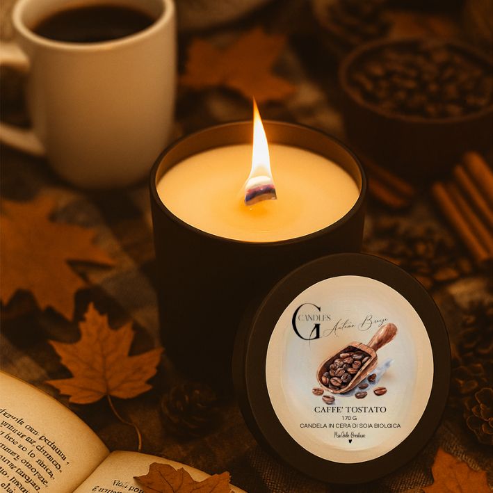 Candela profumata al gusto di caffè tostato in cera di soia biologica colata a mano in barattolo di alluminio nero con stoppino in legno, decorata con illustrazioni con chicchi di caffè, su sfondo con colori caldi, autunnali, tazza di caffè, libro e elementi naturali.