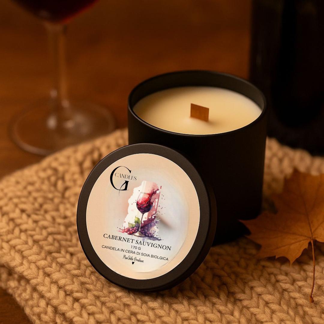 Candela profumata al gusto di vino rosso cabernet sauvignon in cera di soia biologica colata a mano in barattolo di alluminio nero con stoppino in legno, decorata con illustrazioni di bicchiere con liquido rosso scuro, su sfondo con colori caldi, autunnali, elementi naturali e parte di coperta intrecciata.
