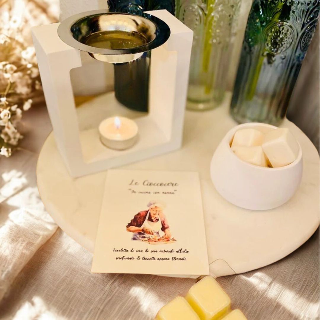 Bruciatore in ceramica bianca con cera da sciogliere profumata ai biscotti, contenitore rotondo, cartoncino illustrato e cubetti di cera su fondo chiaro.