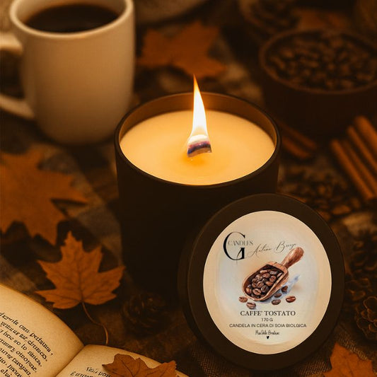Candela profumata al gusto di caffè tostato in cera di soia biologica colata a mano in barattolo di alluminio nero con stoppino in legno, decorata con illustrazioni con chicchi di caffè, su sfondo con colori caldi, autunnali, tazza di caffè, libro e elementi naturali.