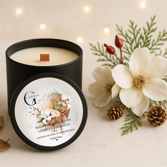 Candela al profumo di magnolia, legno di cedro e vaniglia in cera di soia biologica colata a mano in barattolo di alluminio nero con stoppino in legno, decorata con illustrazioni di elementi naturali, fiori bianchi, pigne, rametti di abete, su sfondo con colori chiari, caldi e lucine natalizie. 