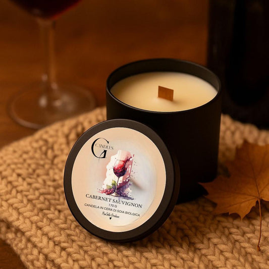 Candela profumata al gusto di vino rosso cabernet sauvignon in cera di soia biologica colata a mano in barattolo di alluminio nero con stoppino in legno, decorata con illustrazioni di bicchiere con liquido rosso scuro, su sfondo con colori caldi, autunnali, elementi naturali e parte di coperta intrecciata.