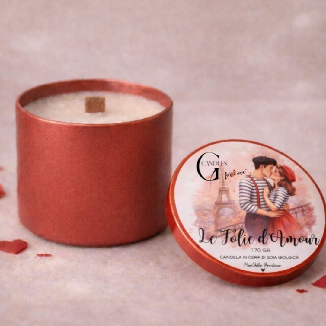Candela in cera di soia bianca al profuma per San Valentino "Le Folie d'Amour" con stoppino in legno in barattolino rosso. Grafica artigianale su coperchio. Piano in legno chiaro e petali rossi come dettagli decorativi.