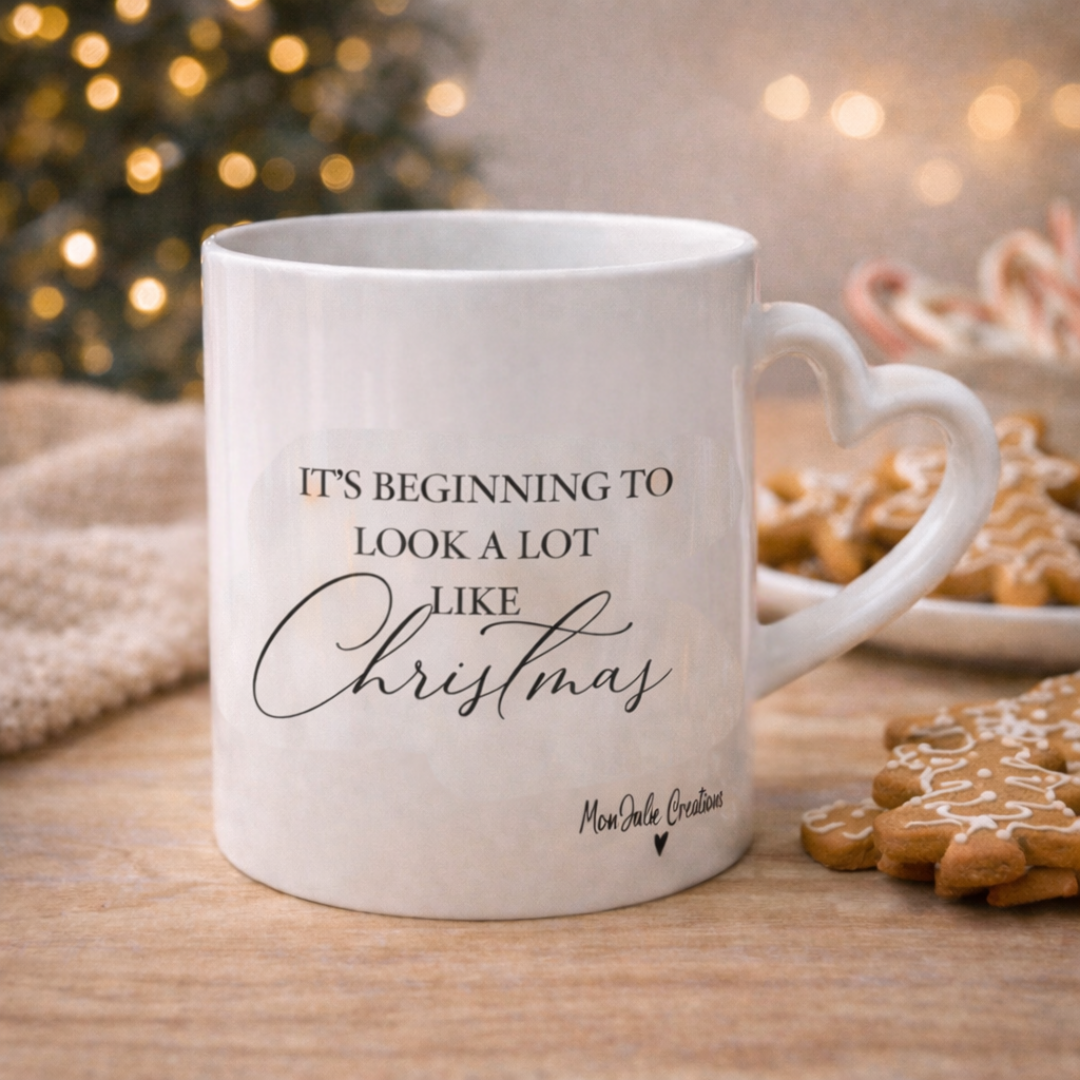 Tazza mug natalizia in ceramica bianca con manico a cuore e scritta, realizzata artigianalmente, su piano in legno chiaro con biscotti e atmosfera di natale con albero e lucine soffuse sullo sfondo.