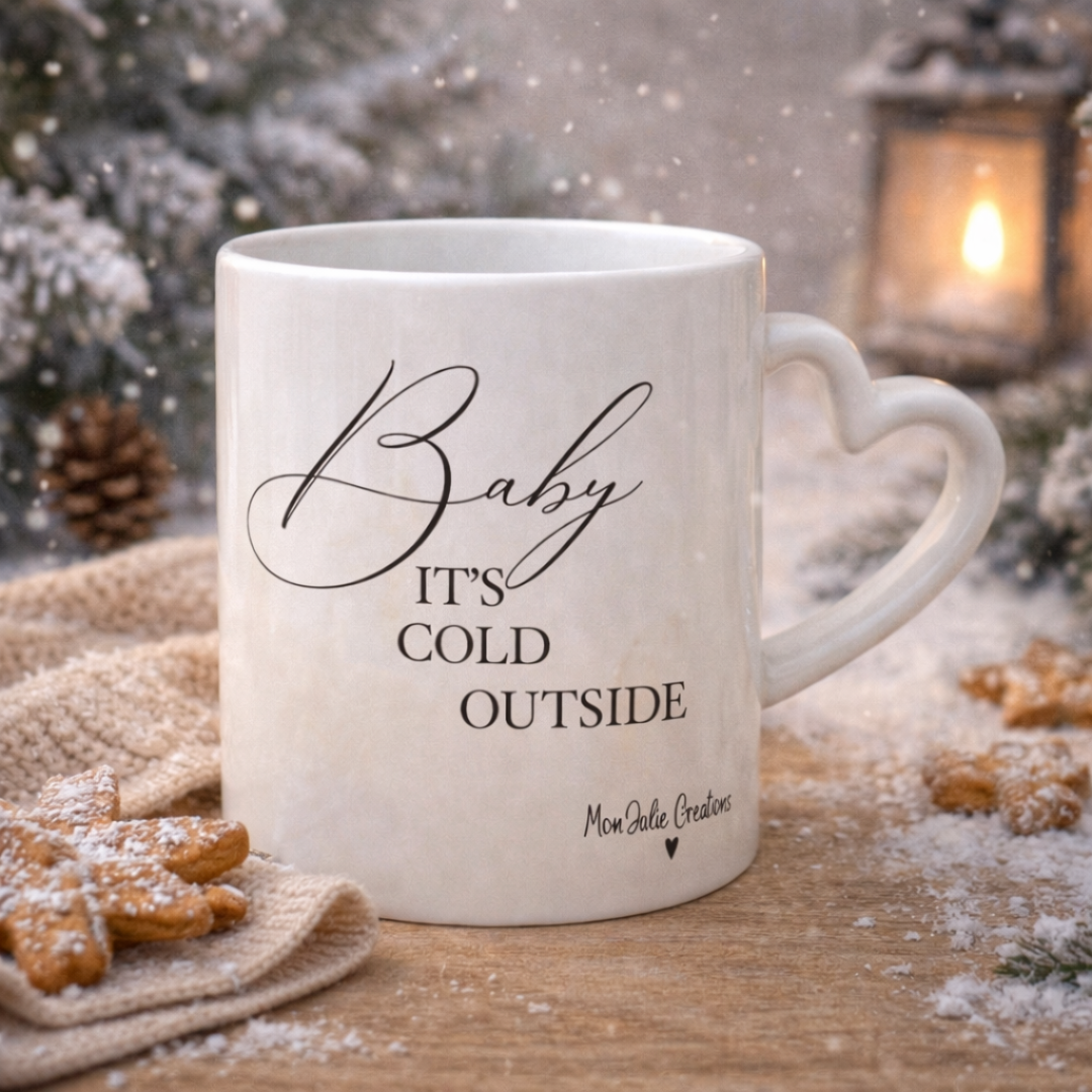 Tazza mug natalizia in ceramica bianca con manico a cuore e scritta, realizzata artigianalmente, su piano in legno chiaro innevato, con biscotti e atmosfera di natale con albero, lucine soffuse, lanterna accesa e neve cadente sullo sfondo.